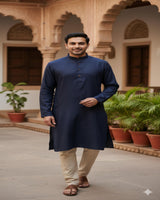 Man wearing a navy kurta with beige pants standing in an outdoor setting with plants and architectural elements.